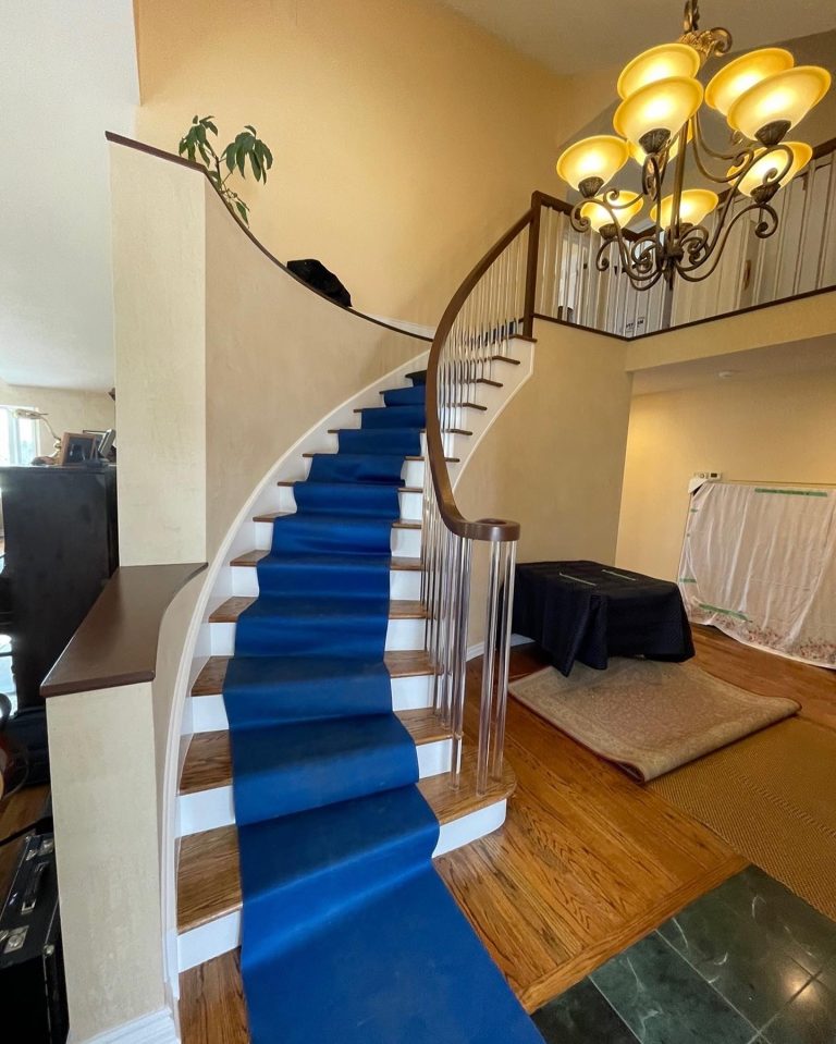 Curved staircase covered by a blue floor runner; a table under a moving blanket and a large piece wrapped in plastic film.