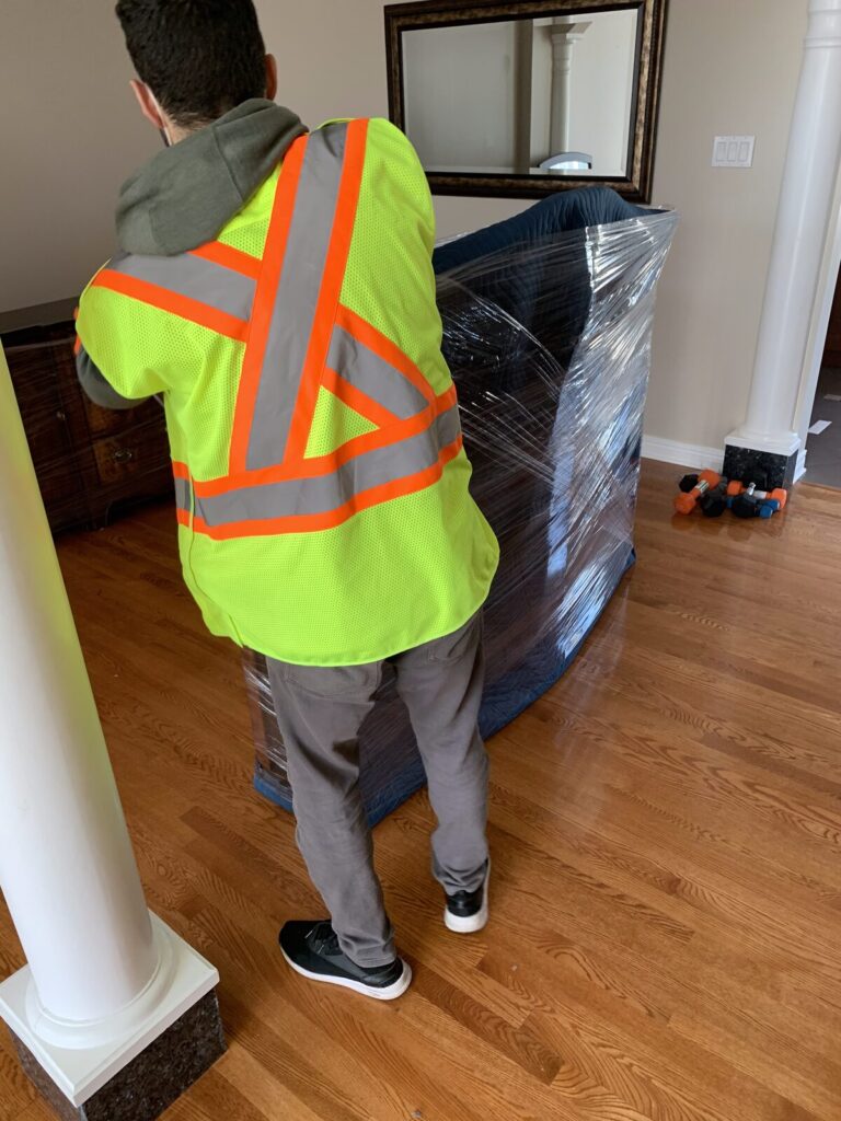 Mover in a hi-vis vest stretch-wrapping a large piece of furniture on a hardwood floor.