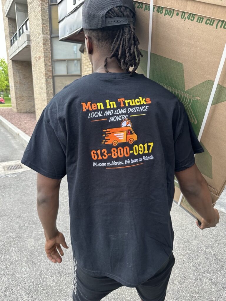 Mover in a Men In Trucks T-shirt carrying a wardrobe box outside a low-rise apartment building.