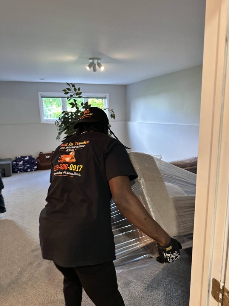 Mover in a Men In Trucks T-shirt and work gloves carrying a shrink-wrapped piece of furniture into a carpeted room.