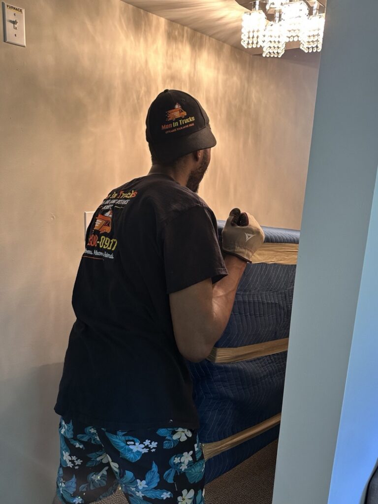 Mover carrying a blanket-wrapped headboard through a room with a chandelier.